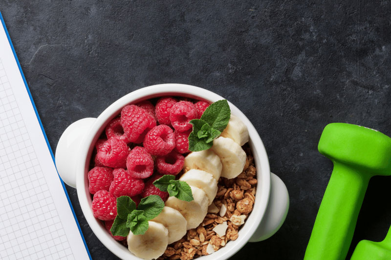 Notebook, bowl of cereal with fruit, and green weights on a table