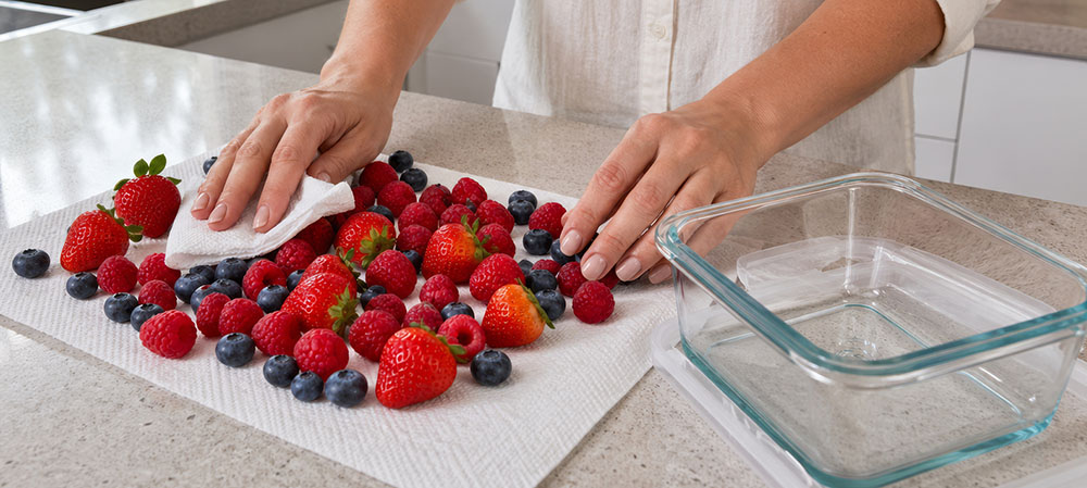 Hands pat dry mixed berries on a paper towel next to an empty glass container on a countertop.