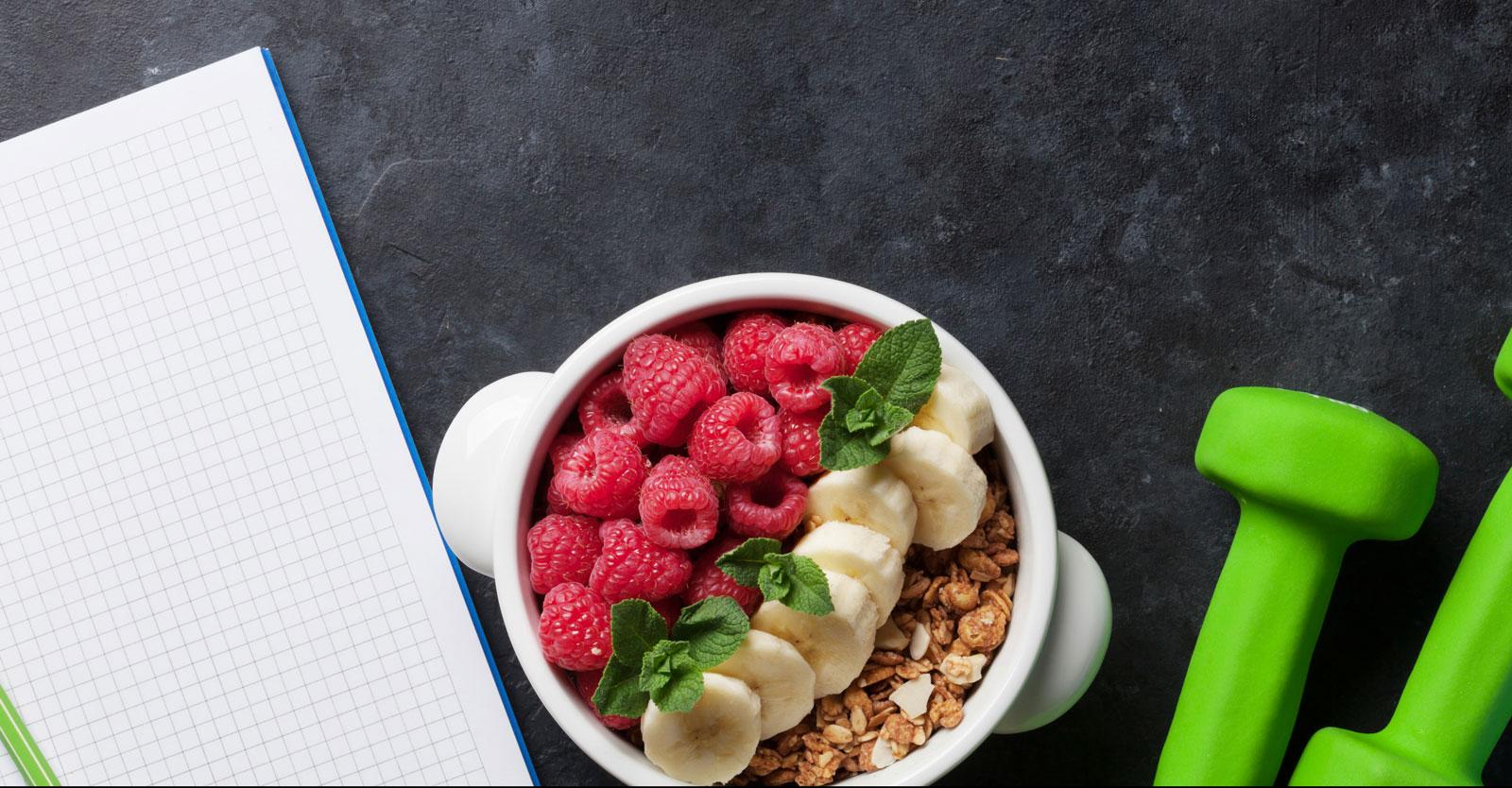 A notebook, a bowl of cereal with fruit, and some green weights on a table.